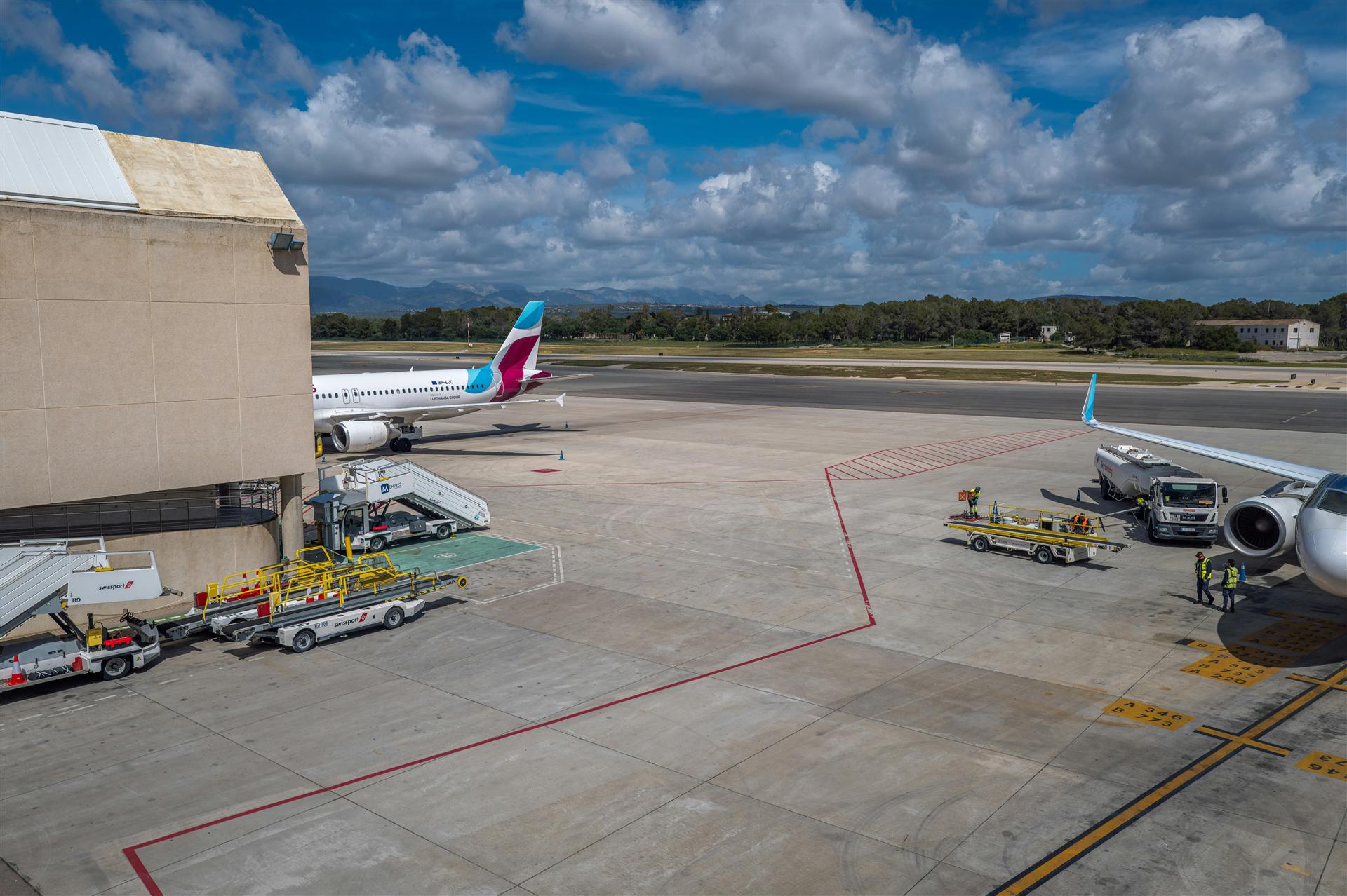 Commercial aircraft parked at an airport gate connected to a passenger boarding bridge, with ground crew on the ramp.